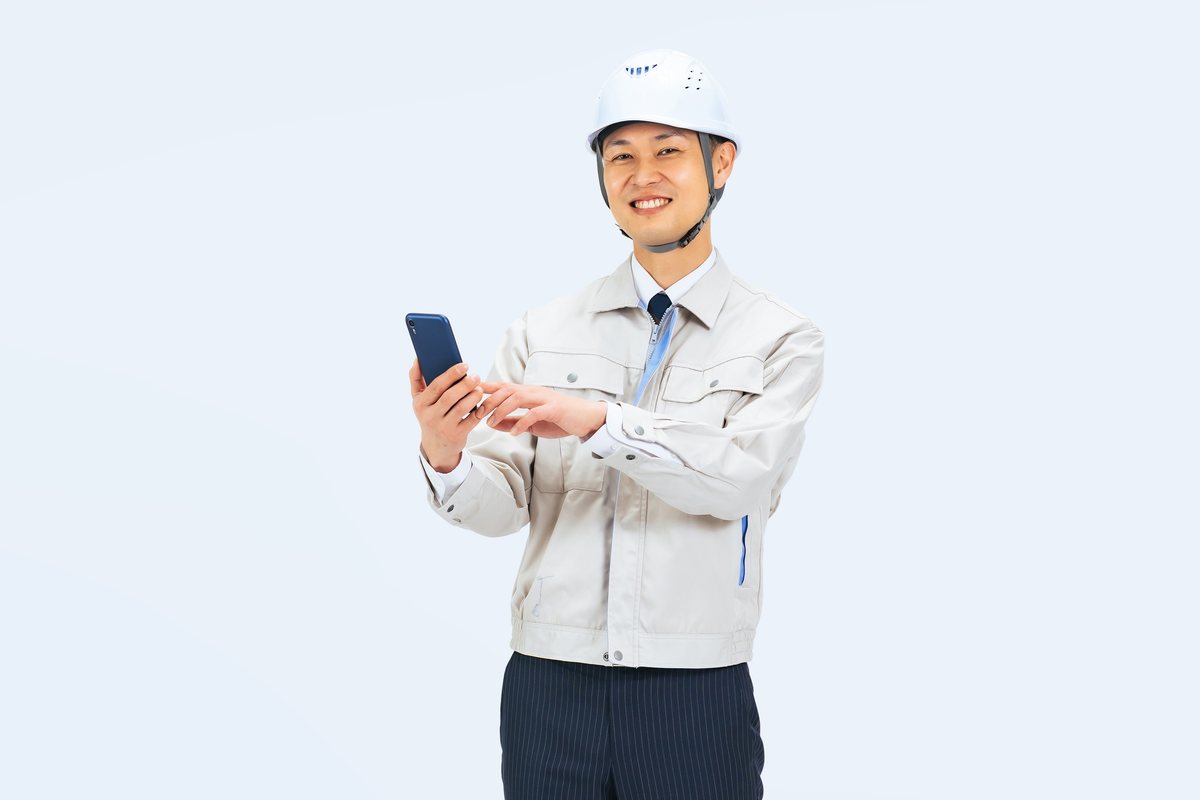 ヘルメットと作業服の男性がスマートフォンを手に笑顔で立っている様子